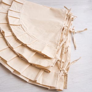 May include: A stack of off-white, ruffled fabric tree skirts with tie closures. The tree skirts have a semi-circular shape with a pleated edge. The fabric appears to be a natural, unbleached cotton or linen material.