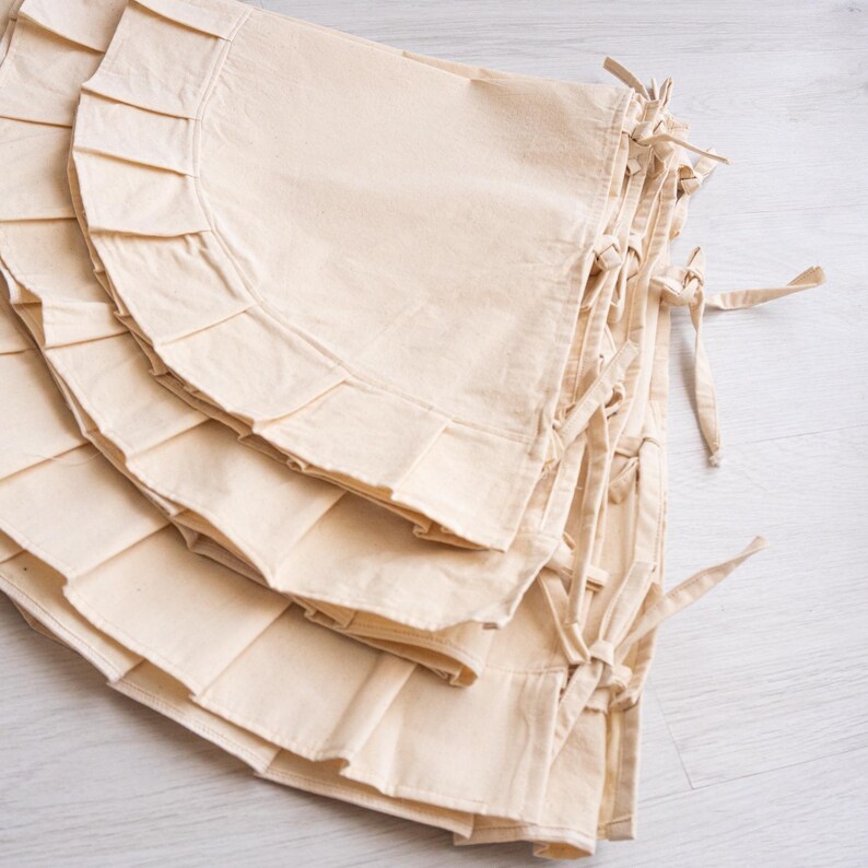 May include: A stack of off-white, ruffled fabric tree skirts with tie closures. The tree skirts have a semi-circular shape with a pleated edge. The fabric appears to be a natural, unbleached cotton or linen material.