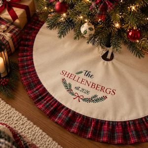 May include: A round cream-colored Christmas tree skirt featuring a red and green plaid ruffled border. The center is embroidered with the text "the SHELLENBERGS est.2026" in red and green, accented by a pine branch and a small red bow design.