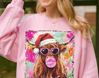 May include: Pink sweatshirt featuring a graphic of a Highland cow wearing a Santa hat and heart-shaped sunglasses, blowing a bubblegum bubble. The cow is set against a colorful, abstract background.