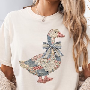 May include: Cream-colored t-shirt featuring a patchwork goose design. The goose has a blue gingham bow, a red and white checkered beak and feet, and a body made of floral and patchwork fabrics. The shirt is a relaxed fit.