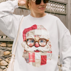 Puede incluir: Sudadera blanca con un diseño de cara de Papá Noel. La cara de Papá Noel está hecha de tela de retazos con un gorro rojo, gafas y barba blanca. El diseño está centrado en la parte delantera de la parte superior de manga larga.