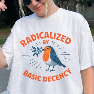 以下が含まれることがあります： オレンジ色の「RADICALIZED BY BASIC DECENCY」の文字が入った白いTシャツ。青とオレンジの鳥のイラストがテキストの下に配置され、小枝を持っています。デザインは放射線状の線で囲まれています。