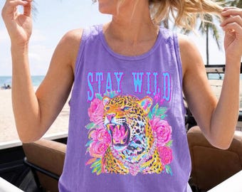 Könnte beinhalten: Ein lila Tanktop mit dem Schriftzug "STAY WILD" in Türkis und Pink. Das Shirt zeigt eine Leoparden-Grafik mit rosa Rosen und einer goldenen Halskette. Das Tanktop ist ärmellos.