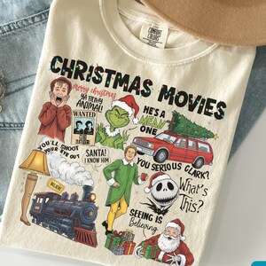 May include: Ivory-colored t-shirt with the text "CHRISTMAS MOVIES" and various cartoon characters and quotes from Christmas movies. The shirt includes images of the Grinch, Santa, and a red station wagon with a Christmas tree.