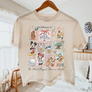 Op de afbeelding: Beige T-shirt met korte mouwen met een kleurrijke afbeelding van Disney-personages en bezienswaardigheden. Het ontwerp bevat Mickey Mouse, Minnie Mouse, Donald Duck en Goofy, samen met de tekst "Walt Disney World" en "2026". Het shirt heeft ook de tekst "The Most Magical Place on Earth".