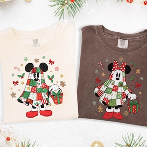 May include: Two t-shirts, one cream and one brown, featuring a Christmas-themed cartoon character. The character is dressed in a patchwork quilt costume, holding a gift basket, and surrounded by festive holiday graphics.