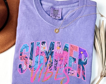 May include: A lilac Comfort Colors t-shirt with the words "SUMMER VIBES" in a floral design. A gold necklace with a pendant rests on the shirt. A brown hat and dried plants are in the background.