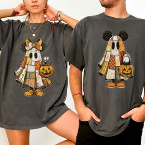 Peut inclure: T-shirts gris foncé avec un motif de fantôme d'Halloween. Le fantôme est un patchwork de différentes couleurs et motifs, avec des oreilles de Mickey Mouse et tenant une lanterne citrouille. Les chemises sont surdimensionnées et à manches courtes.