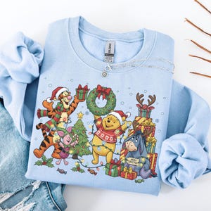 May include: A light blue sweatshirt featuring a festive Christmas design with Winnie the Pooh characters. The graphic includes Tigger, Piglet, Winnie the Pooh, and Eeyore, surrounded by Christmas lights, gifts, and a Christmas tree. A silver necklace is draped on the sweatshirt.