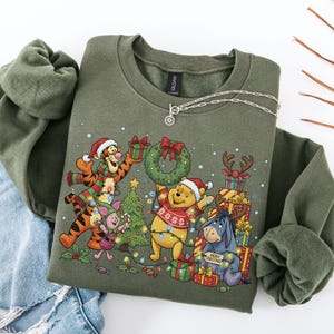 May include: An olive green sweatshirt featuring a festive Christmas design with Winnie the Pooh and friends. The graphic includes characters wearing Santa hats, holding gifts, and surrounded by Christmas lights and a wreath. A silver necklace is draped across the sweatshirt.