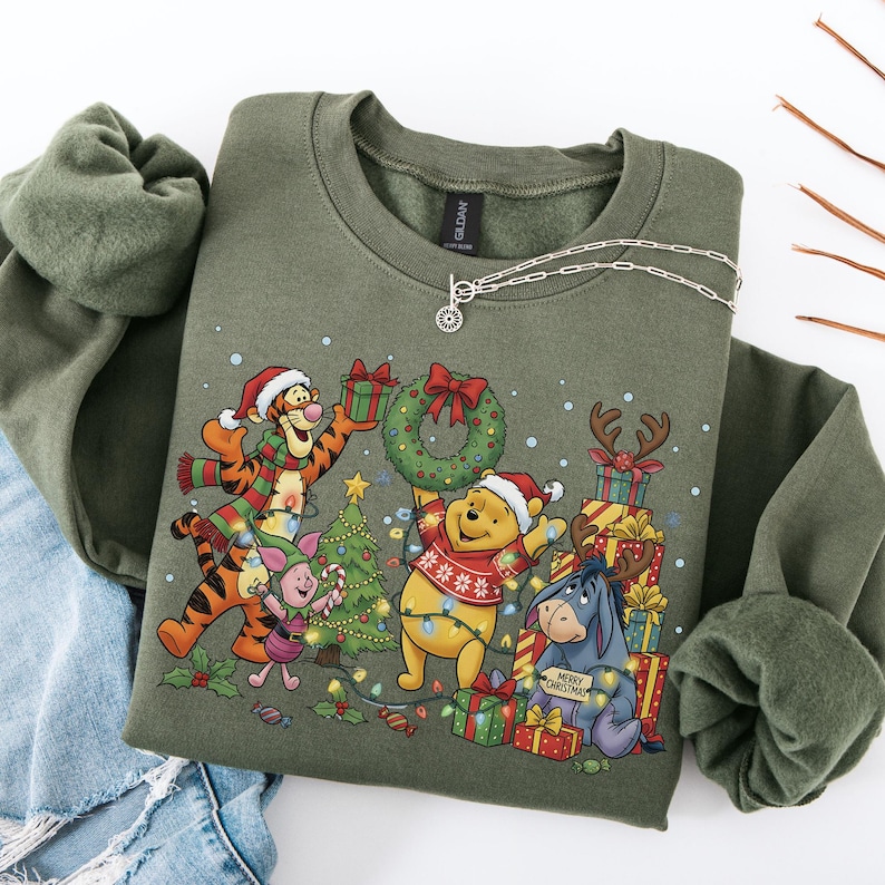 May include: An olive green sweatshirt featuring a festive Christmas design with Winnie the Pooh and friends. The graphic includes characters wearing Santa hats, holding gifts, and surrounded by Christmas lights and a wreath. A silver necklace is draped across the sweatshirt.