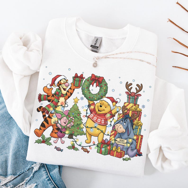 May include: White sweatshirt featuring a festive graphic of Winnie the Pooh and friends celebrating Christmas. The design includes characters like Tigger, Piglet, and Eeyore, surrounded by presents, a Christmas tree, and holiday lights. A silver necklace is draped on the sweatshirt.