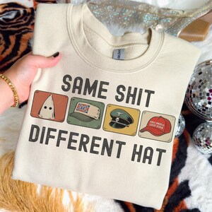 Peut inclure: Sweat-shirt crème avec l'inscription "SAME SHIT DIFFERENT HAT" au-dessus de quatre images de chapeaux. Les chapeaux comprennent une cagoule blanche, un chapeau confédéré, un chapeau militaire et un chapeau rouge "Make America Great Again".