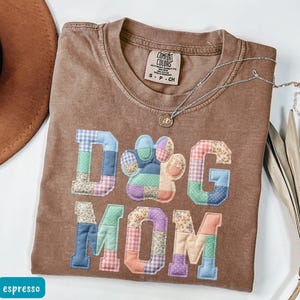 May include: Espresso-coloured t-shirt featuring the text "DOG MOM" in patchwork lettering and a paw print. The letters and paw print are made of various patterned fabrics in shades of blue, green, pink, and purple. A silver necklace with a pendant is also visible.