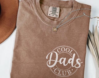 May include: A brown Comfort Colors t-shirt with the text "COOL Dads CLUB" in a white circular design. A silver necklace with a small pendant is draped across the shirt. The shirt is size small.