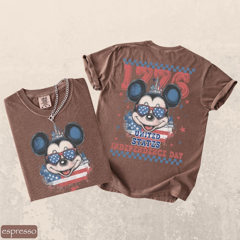 Puede incluir: Dos camisetas marrones con un dise&ntilde;o de Mickey Mouse con gafas de sol y un dise&ntilde;o de bandera americana. Las camisetas muestran el texto "1776" y "United States Independence Day". Una camiseta tiene una cadena plateada.