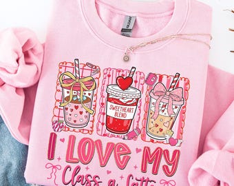 Puede incluir: Sudadera rosa claro con un gráfico de tres tazas de café con detalles de corazón y lazos. El texto "I Love My Class a Latte" está impreso en rosa. También se ve un delicado collar con un colgante.