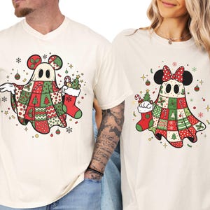 May include: Two cream-colored t-shirts featuring a cartoon ghost design with Mickey Mouse ears. The ghosts are wearing patchwork cloaks in red, green, and white, holding Christmas stockings and trees. Festive stars and ornaments surround the ghosts.