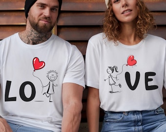 Puede incluir: Camisetas blancas con la palabra "LOVE" en letras negras. La "O" se reemplaza con un muñeco de palo que sostiene un globo rojo en forma de corazón. La otra camiseta tiene la "VE" con un muñeco de palo y un globo rojo.