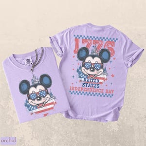 Puede incluir: Dos camisetas de color lila claro con un gr&aacute;fico de Mickey Mouse con gafas de sol con un dise&ntilde;o patri&oacute;tico. Las camisetas tienen el texto "1776" y "United States Independence Day". Una camiseta tiene una cadena plateada.