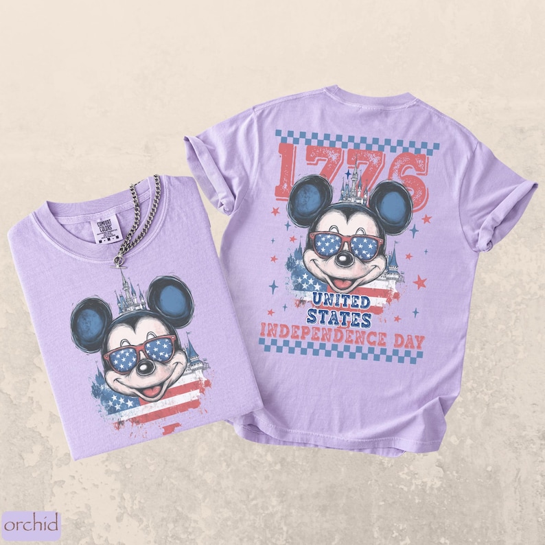 Puede incluir: Dos camisetas de color lila claro con un gr&aacute;fico de Mickey Mouse con gafas de sol con un dise&ntilde;o patri&oacute;tico. Las camisetas tienen el texto "1776" y "United States Independence Day". Una camiseta tiene una cadena plateada.