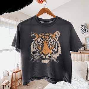 May include: A charcoal gray short-sleeve t-shirt featuring a large, centered graphic print of a tiger face. The tiger illustration is rendered in shades of orange, cream, and black with a distressed, weathered texture across the front of the shirt.