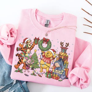 May include: Pink sweatshirt featuring Winnie the Pooh, Tigger, Piglet, and Eeyore in Christmas attire. The graphic includes a Christmas tree, presents, and a wreath. The characters are surrounded by Christmas lights. A necklace is draped across the sweatshirt.