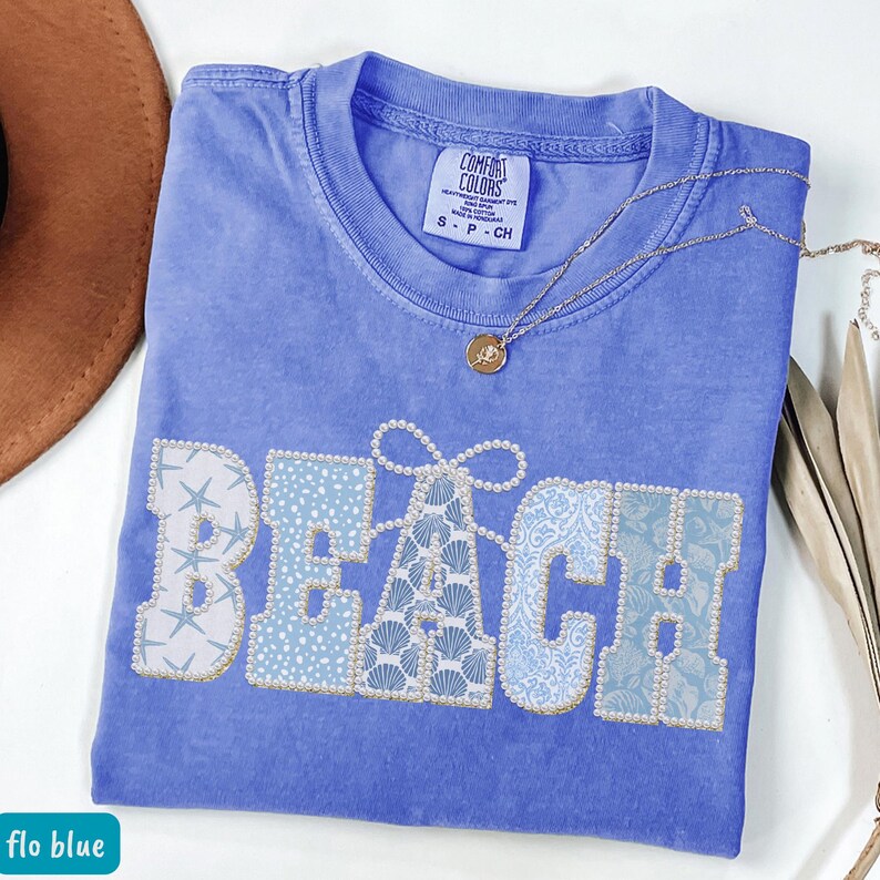 May include: A blue Comfort Colors t-shirt with the word "BEACH" embroidered in a mix of patterns and embellished with pearls. A gold necklace with a pendant rests on the shirt. The shirt is labeled "flo blue".