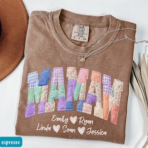May include: Espresso-colored t-shirt with the word "MAMA" in patchwork letters. The letters are in various colors and patterns. Below the word are names with heart symbols. A silver necklace is draped across the shirt.