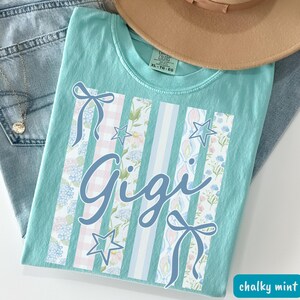 May include: A chalky mint green t-shirt with the name "Gigi" in a cursive font. The shirt features vertical stripes with floral and gingham patterns, bows, and stars. A tan hat and blue jeans are in the background.