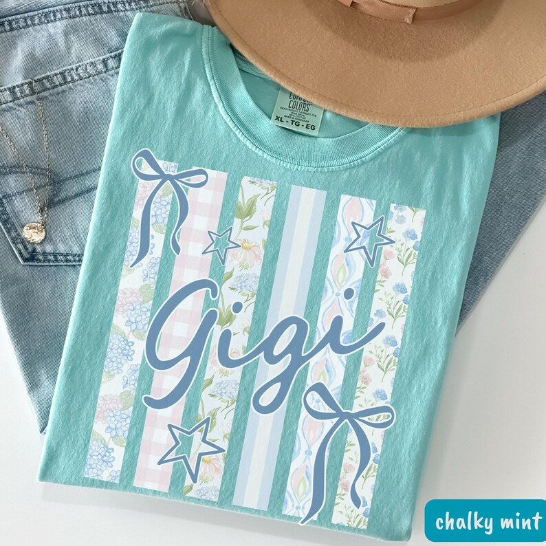 May include: A chalky mint green t-shirt with the name "Gigi" in a cursive font. The shirt features vertical stripes with floral and gingham patterns, bows, and stars. A tan hat and blue jeans are in the background.