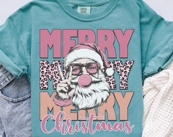 May include: Seafoam green Comfort Colors t-shirt featuring a graphic of Santa Claus wearing sunglasses and blowing a bubblegum bubble. The text "MERRY CHRISTMAS" is displayed in pink and orange.