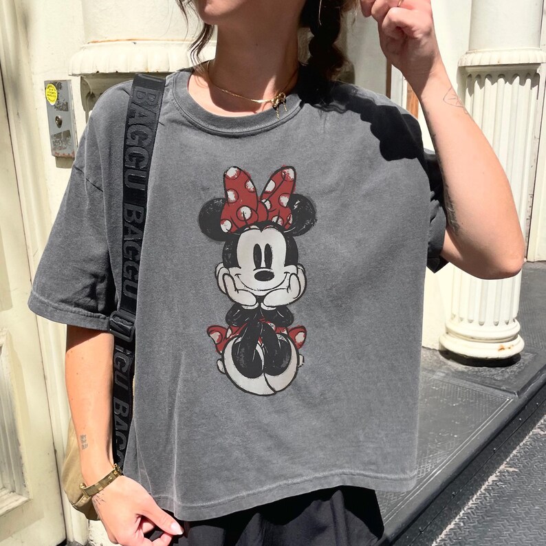 Pode incluir: Uma camiseta cinza de manga curta com um gr&aacute;fico da Minnie Mouse. Minnie &eacute; retratada com um la&ccedil;o vermelho de bolinhas e em uma pose sentada. A camiseta tem uma apar&ecirc;ncia ligeiramente desbotada.