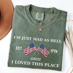 May include: Olive green t-shirt with the text "I'M JUST MAD AS HELL" and "CAUSE I LOVED THIS PLACE". The shirt features two American flags and the year 1776. A gold necklace with a pendant is draped across the shirt.