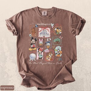 Op de afbeelding: Espresso-kleurig T-shirt met een collage van Disney-personages en bezienswaardigheden. Het ontwerp bevat Mickey Mouse, Minnie Mouse, Donald Duck en Goofy, samen met de tekst "Walt Disney World" en "2026". Het shirt heeft ook de tekst "The Most Magical Place on Earth".