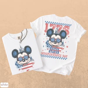 Puede incluir: Dos camisetas blancas con un dise&ntilde;o de Mickey Mouse con gafas de sol de dise&ntilde;o patri&oacute;tico. Una camiseta muestra el dise&ntilde;o en la parte delantera, la otra en la trasera, con el texto "1776" y "United States Independence Day".
