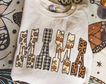 Pode incluir: Um moletom bege com a palavra "MAMA" em uma fonte decorativa. As letras são estampadas com estampa de leopardo, xadrez preto e branco e abóboras. O moletom está dobrado e é segurado por mãos.