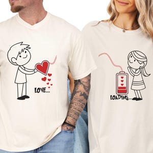 May include: Two cream-coloured t-shirts with black line art. One shirt features a stick figure holding a red heart with the word "LOVE..." below. The other shirt shows a stick figure with a battery graphic and the word "LOADING..."