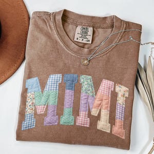 Può includere: Una maglietta marrone Comfort Colors con il nome "MIMI" applicato sul davanti in tessuto patchwork. Le lettere patchwork sono in vari colori e motivi pastello. Una collana d'argento con un piccolo ciondolo è drappeggiata sulla maglietta.