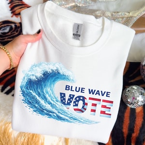 May include: White sweatshirt featuring a blue wave graphic and the words "BLUE WAVE VOTE" in navy blue and red. The word "VOTE" incorporates a patriotic stars and stripes design. The sweatshirt is held by a person.