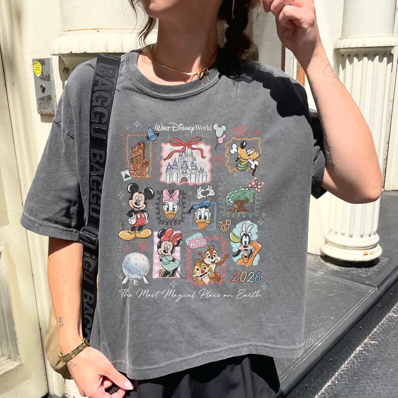 Op de afbeelding: Grijs T-shirt met een Disney-ontwerp. De print bevat verschillende personages zoals Mickey Mouse, Donald Duck en Goofy, samen met de tekst "Walt Disney World" en het jaar "2026". De tekst "The Most Magical Place on Earth" staat ook op het shirt.