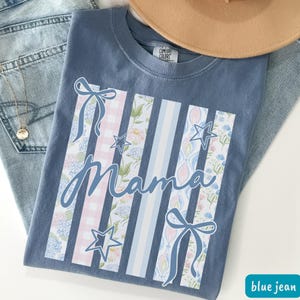 May include: A blue jean Comfort Colors t-shirt with the word "Mama" in script. The design includes vertical stripes with floral, gingham, and star patterns, and bows. A gold necklace and a tan hat are also visible.