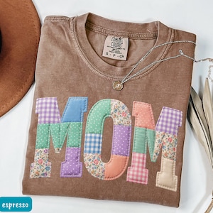 May include: Espresso-colored t-shirt with the word "MOM" appliqued in colorful patchwork fabric. The shirt has a crew neck and short sleeves. A silver necklace with a pendant is draped across the shirt. The shirt is labeled "Comfort Colors".