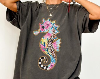 May include: Dark gray t-shirt featuring a colorful seahorse design. The seahorse is decorated with floral, leopard print, and checkered patterns, outlined with gold glitter. The shirt has short sleeves and a relaxed fit.