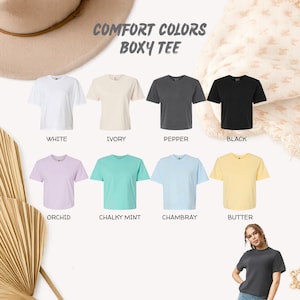 Op de afbeelding: Een display van acht boxy T-shirts in verschillende kleuren: wit, ivoor, peper, zwart, orchidee, krijtmunt, chambray en boter. De tekst "COMFORT COLORS BOXY TEE" staat bovenaan. Een vrouw draagt een peperkleurig T-shirt.