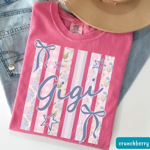 May include: A pink Comfort Colors t-shirt with the name "Gigi" in a cursive font. The shirt features vertical stripes with floral and star patterns, and bows. The word "crunchberry" is in a teal box in the bottom right corner.