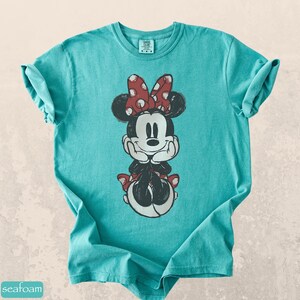Pode incluir: Uma camiseta turquesa com um gr&aacute;fico desgastado da Minnie Mouse. Minnie &eacute; retratada com um la&ccedil;o vermelho de bolinhas, orelhas pretas e uma roupa preta e branca. A camiseta tem mangas enroladas e gola redonda.