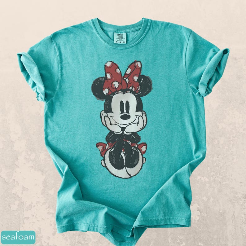 Pode incluir: Uma camiseta turquesa com um gr&aacute;fico desgastado da Minnie Mouse. Minnie &eacute; retratada com um la&ccedil;o vermelho de bolinhas, orelhas pretas e uma roupa preta e branca. A camiseta tem mangas enroladas e gola redonda.