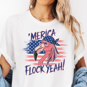 Könnte beinhalten: Weißes T-Shirt mit einem Flamingo, der eine Sonnenbrille und ein Bandana trägt, vor dem Hintergrund der amerikanischen Flagge. Der Text "'Merica Flock Yeah!" ist in patriotischen Farben gehalten.